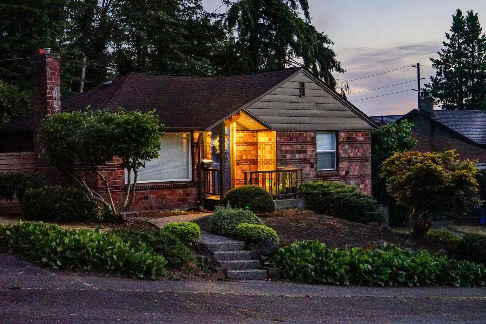 Brick rambler with a lit porch and landscaped yard at dusk
