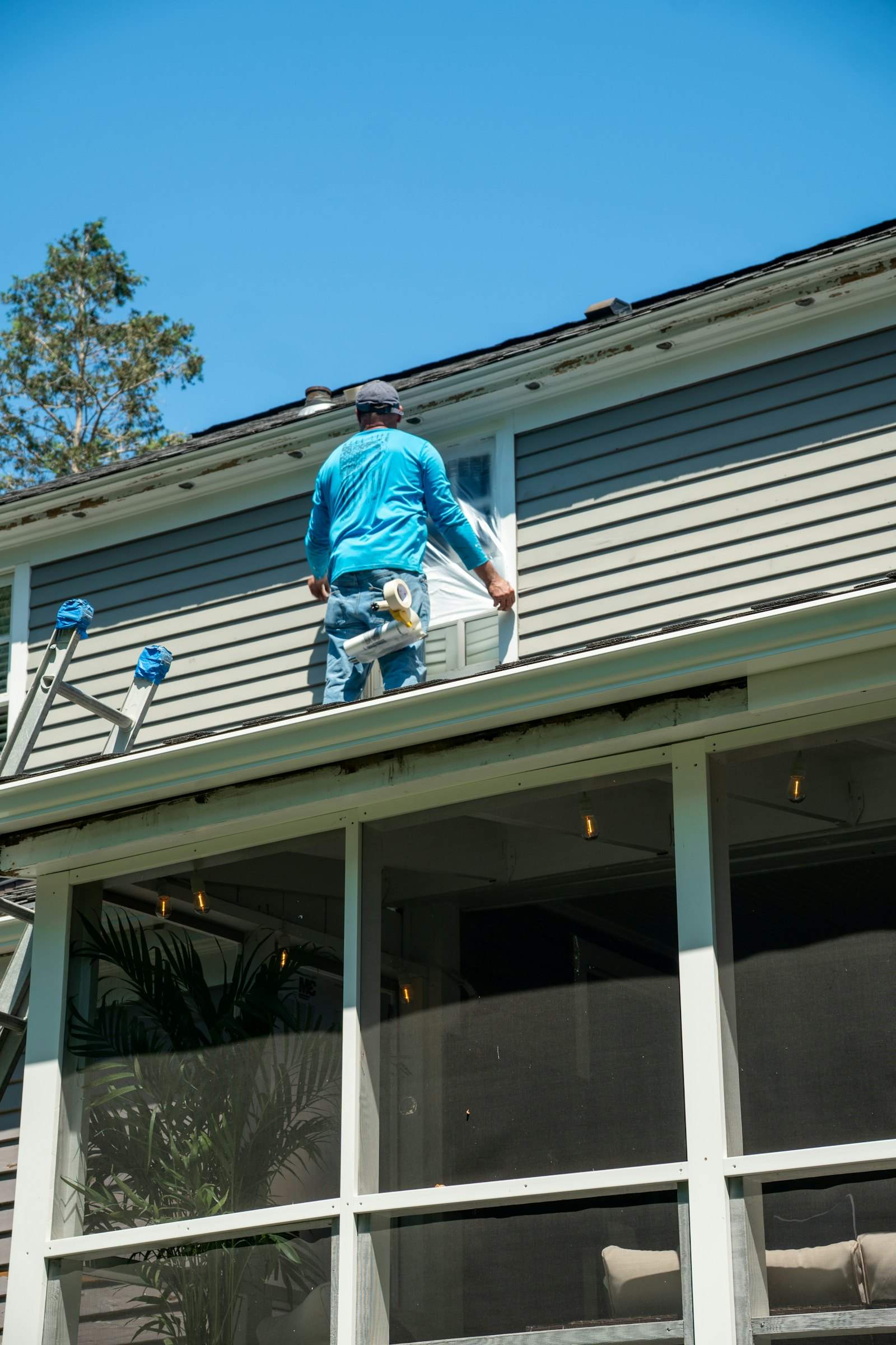 Worker on a porch roof taping plastic over a window before exterior work