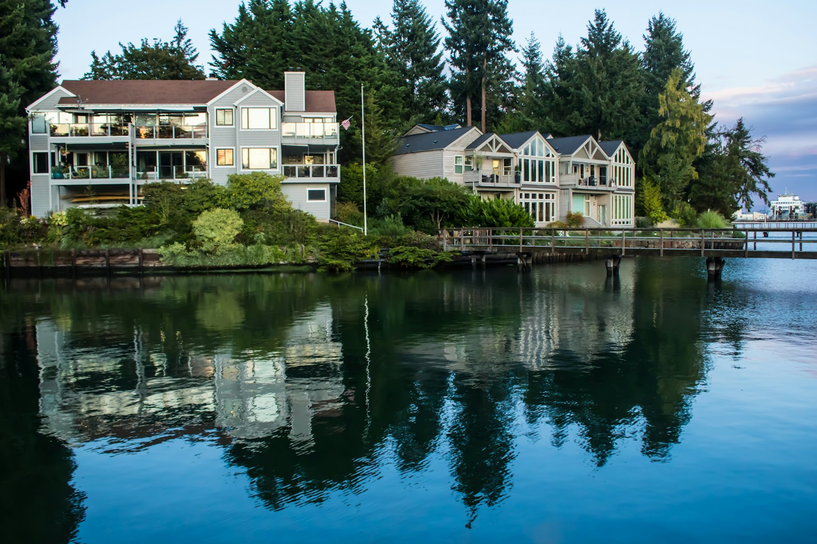 Lakefront homes with large windows reflected in calm water, surrounded by evergreens