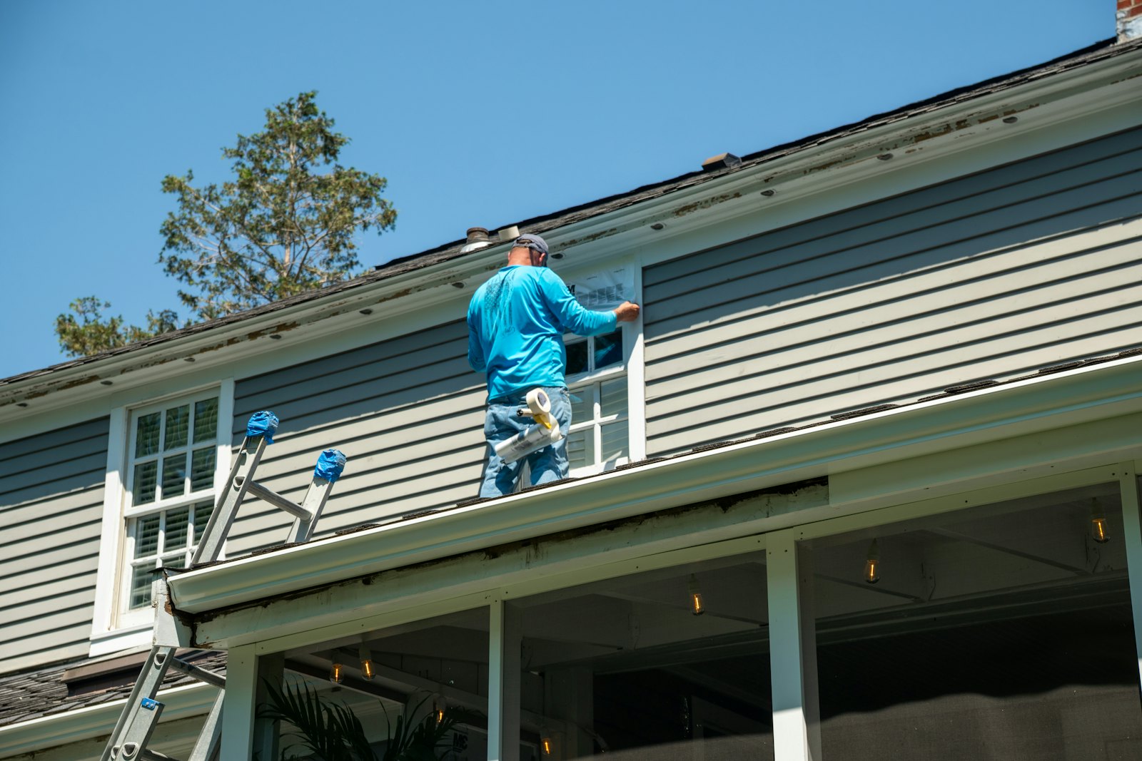 Worker on a porch roof sealing a window surrounded by gray lap siding